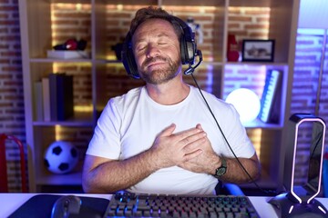 Middle age man with beard playing video games wearing headphones smiling with hands on chest with closed eyes and grateful gesture on face. health concept.
