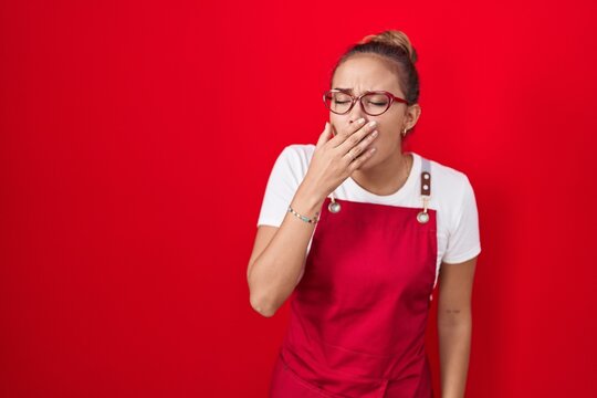 Young hispanic woman wearing waitress apron over red background bored yawning tired covering mouth with hand. restless and sleepiness.