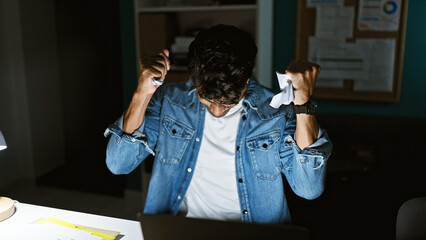 Frustrated hispanic man crumples paper in a dimly lit office, showcasing stress, work, and...