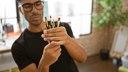 A focused young hispanic man examines paintbrushes in a creative studio setting.