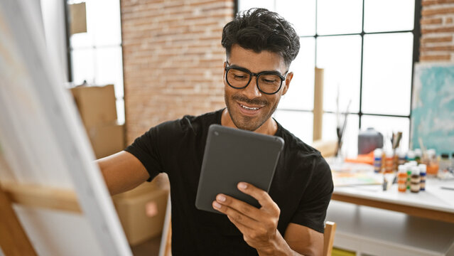 Smiling young hispanic man with beard using tablet in art studio.