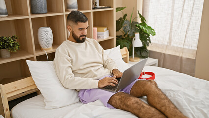 Bearded man using laptop in a modern bedroom setting, reflecting a blend of leisure and productivity.