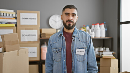 A confident young man with a beard stands as a volunteer in a cluttered storage room, boxed...