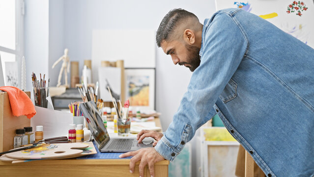 Handsome bearded man in casual denim attire working on a laptop in a vibrant art studio.