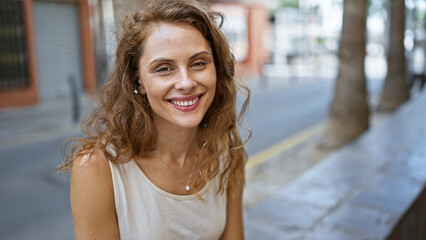 A smiling young woman with wireless earphones enjoys a sunny urban streetscape in the background.