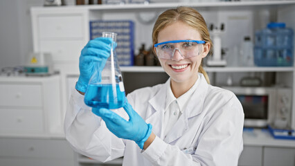 A smiling young woman scientist examining a blue liquid in a flask in a laboratory setting.