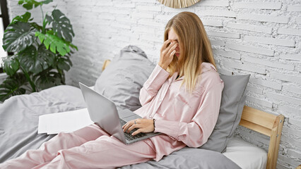 A stressed woman in pajamas working on a laptop in bed, rubbing her eyes, with a sheet of paper...