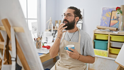 A thoughtful bearded man with glasses and an apron holding a mug in an art studio full of paintings...