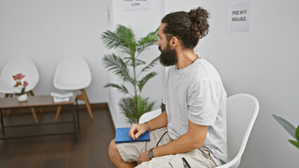 Handsome hispanic man with a beard sitting in a modern waiting room holding a blue notebook