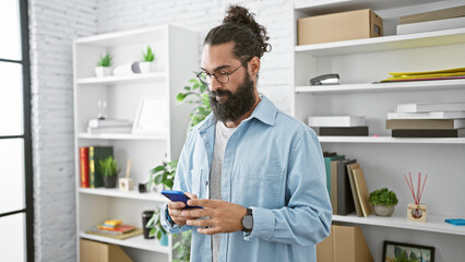 A bearded hispanic man in casual attire focused on his smartphone in a bright modern office.