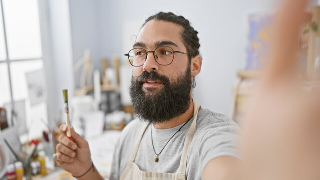 Hispanic man with beard taking a selfie in an art studio, surrounded by painting supplies.
