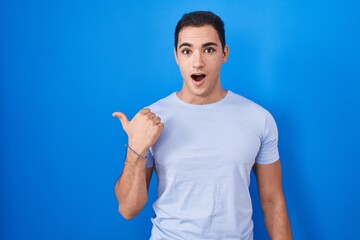 Young hispanic man standing over blue background surprised pointing with hand finger to the side, open mouth amazed expression.