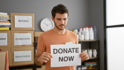 Hispanic man holding a 'donate now' sign in a warehouse filled with donation boxes.