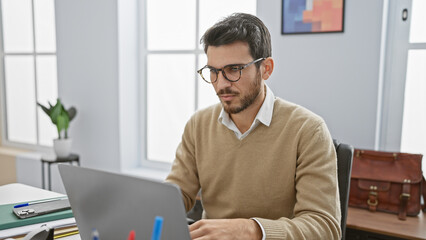 Handsome hispanic man with beard wearing glasses working on laptop in modern office setting