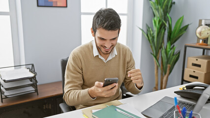 Handsome young hispanic man excitedly looks at smartphone in a modern office setting
