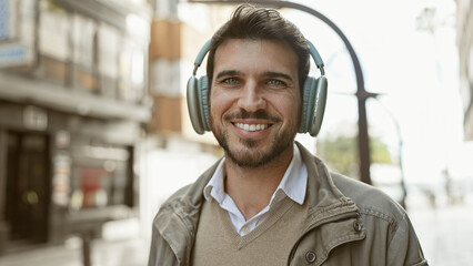 A smiling young hispanic man with a beard enjoys music on headphones while standing on a sunny...
