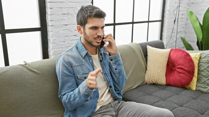 A handsome young hispanic man in casual denim attire talks on a smartphone while seated on a sofa...