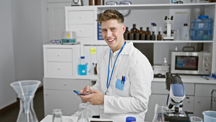 Fototapeta na wymiar Cheerful young caucasian man, a skilled scientist, working with a radiant smile and confidence, expertly texting data on his smartphone at the bustling laboratory.