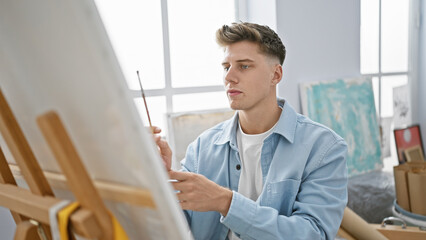 Naklejka na ściany i meble Handsome bearded young caucasian man, an artist, concentrated and immersed in his craft, sits drawing in art class at the university studio, creating magic on canvas.