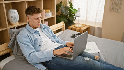 Handsome young caucasian man, sitting in bed, intensely studying on his laptop in his tranquil bedroom morning scene, focused on his study.