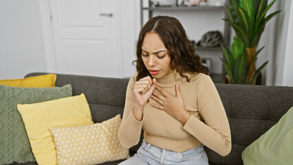 Indoor portrait of a young woman coughing on a couch with colorful pillows around her, seemingly...
