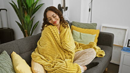 A content woman wrapped in a yellow blanket relaxes on a grey sofa in a modern living room with...
