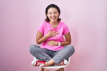 Hispanic young woman sitting on chair over pink background smiling and laughing hard out loud...