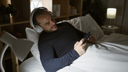 Hispanic man with beard enjoying music on headphones while holding smartphone in bedroom at night.