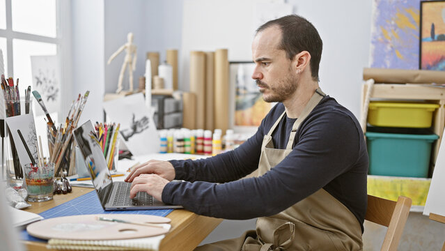 A bald man with a beard wearing an apron uses a laptop in a sunlit art studio full of painting supplies.