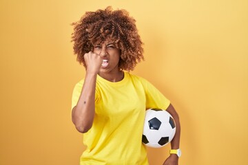 Young hispanic woman with curly hair holding football ball annoyed and frustrated shouting with...