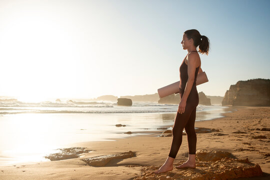 fit sporty woman holding yoga mat, standing on beach at sunrise