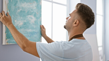 Handsome man hanging blue abstract painting on white wall indoors, showcasing contemporary art.