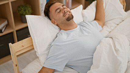 Obraz premium Handsome hispanic man resting in a bedroom with white bedding and a peaceful expression