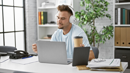 Hispanic man with beard wearing headphones concentrates on laptop in modern office.
