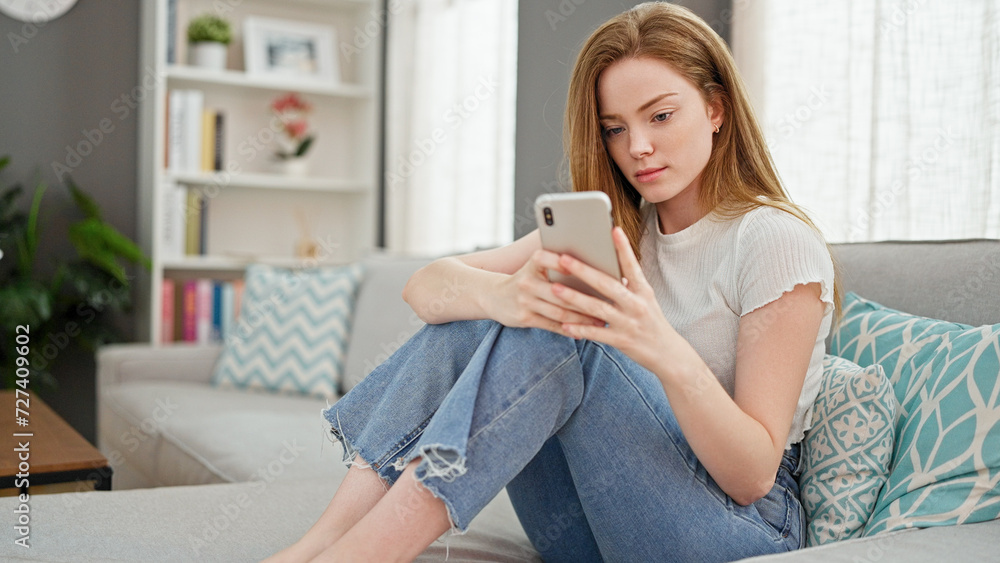 Poster young blonde woman using smartphone sitting on sofa at home