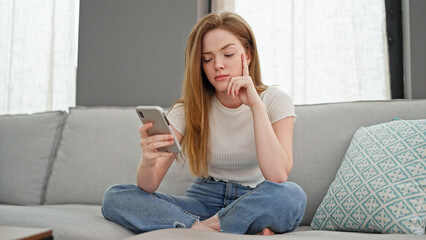 Young blonde woman using smartphone looking upset at home