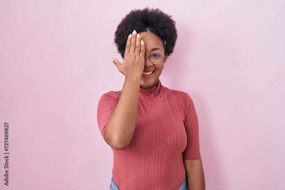 Sticker Beautiful african woman with curly hair standing over pink background covering one eye with hand, confident smile on face and surprise emotion.