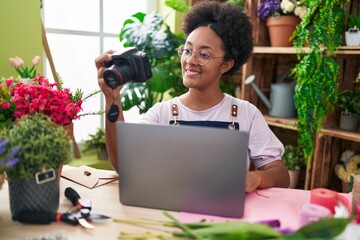 African american woman florist using laptop holding professional camera at flower shop