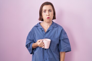 Middle age hispanic woman drinking a cup coffee puffing cheeks with funny face. mouth inflated with...
