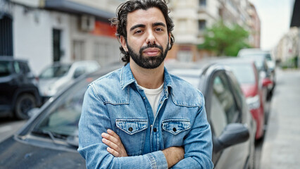 Young hispanic man standing by car with arms crossed gesture and serious face at street
