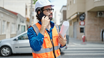 Young hispanic man architect holding blueprints talking on smartphone at construction place