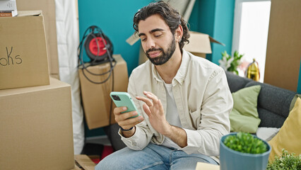 Young hispanic man using smartphone sitting on sofa at new home