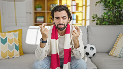 Young hispanic man watching soccer match crossing fingers at home