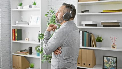 Pensive senior hispanic man with beard and grey hair wearing headphones in a modern office setting.