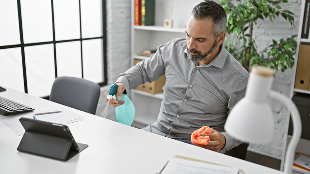 Mature hispanic man disinfecting office desk with spray bottle and cloth