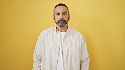 Handsome senior hispanic man with grey beard stands against an isolated yellow wall background,...