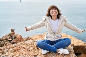 Middle age woman smiling confident sitting with arms open at seaside