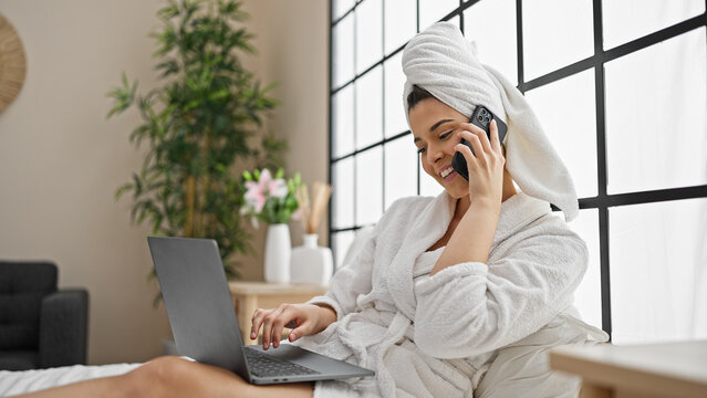 Young beautiful hispanic woman wearing bathrobe using laptop talking on smartphone at bedroom - Powered by Adobe