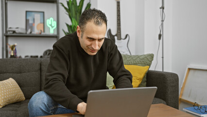 A focused man works on his laptop in a cozy living room decorated with plants and a neon sign.