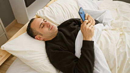 A hispanic man relaxes on a bed with white sheets, casually browsing his smartphone in a cozy bedroom setting.